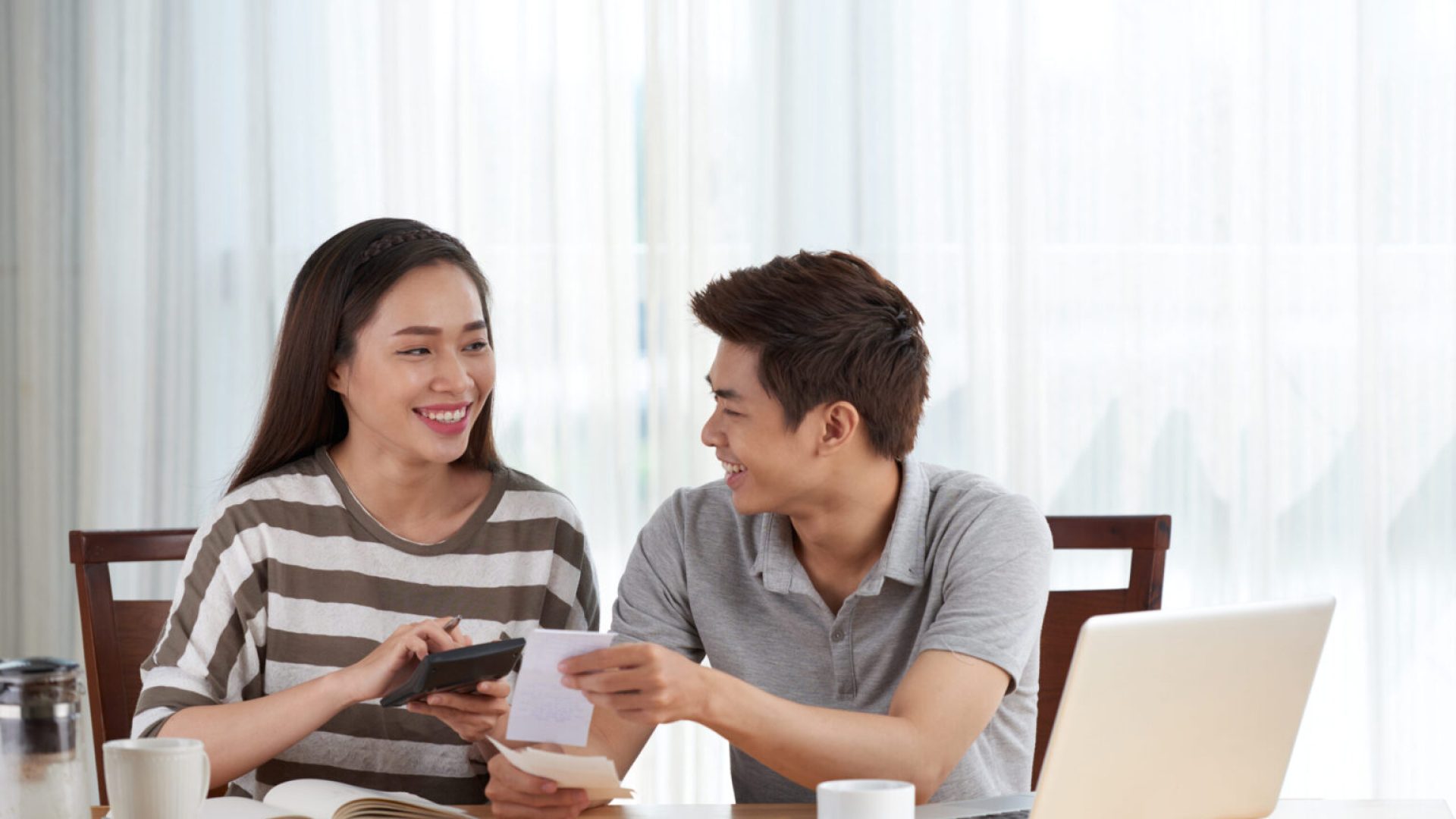 Cheerful pretty woman and her handsome husband managing family budget while sitting at wooden table, interior of living room on background